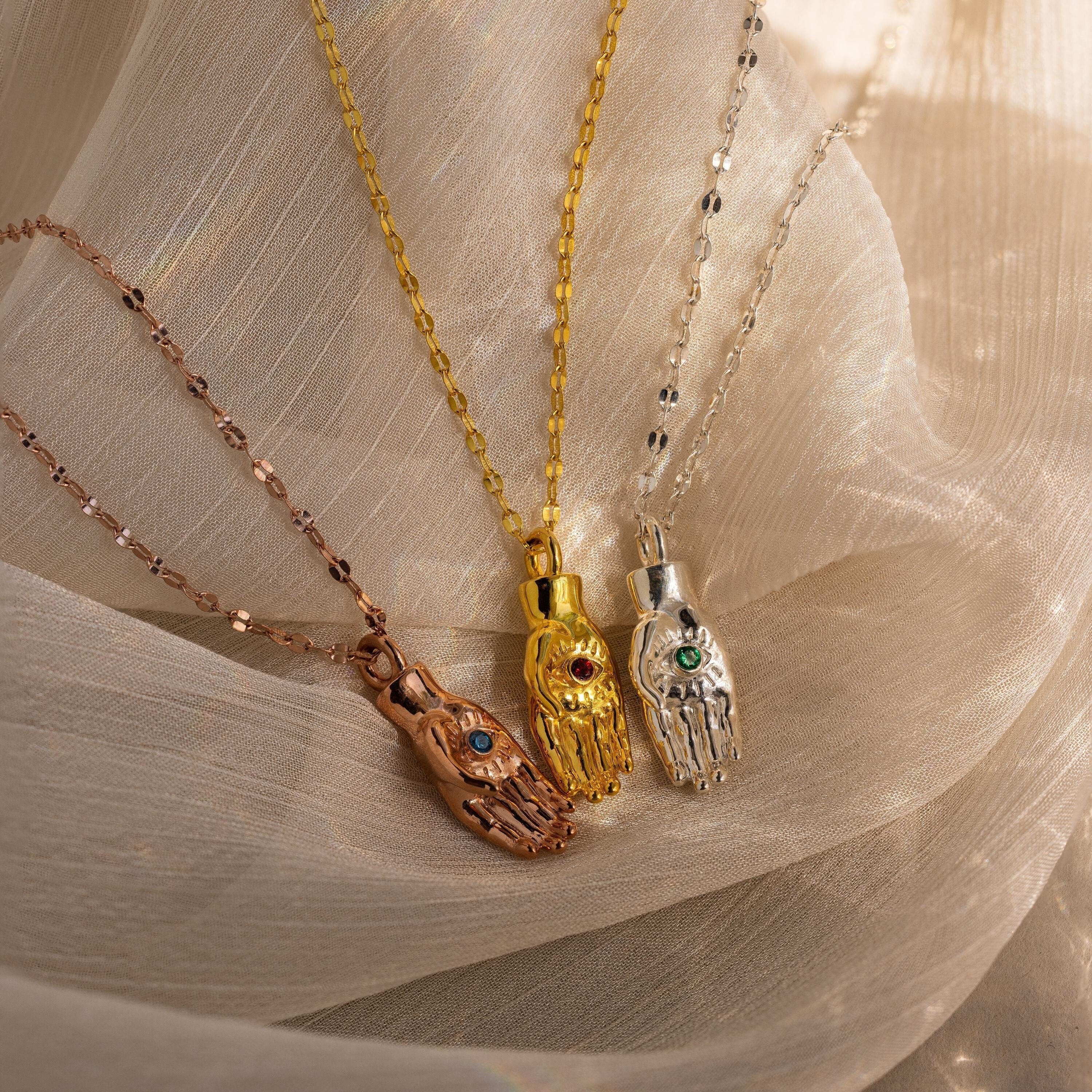 Three Evil Eye Birthstone Hand Necklaces—rose gold, gold, and silver—on matching chains rest elegantly over beige fabric.