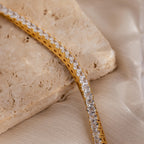 Princess Diamond Tennis Bracelet in 18K Gold displayed on a beige stone and silky fabric background.