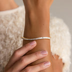 A hand with nude nails touches a wrist adorned with the Princess Diamond Tennis Bracelet in 18K Gold, radiating sparkling elegance.