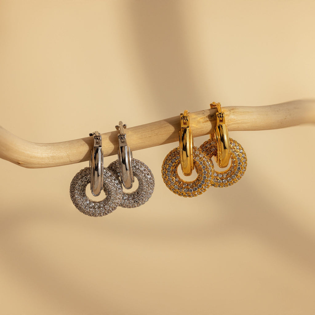 The Duo Pave Link Hoops, featuring bold silver and gold pairs, hang elegantly on a curved branch against a beige background.