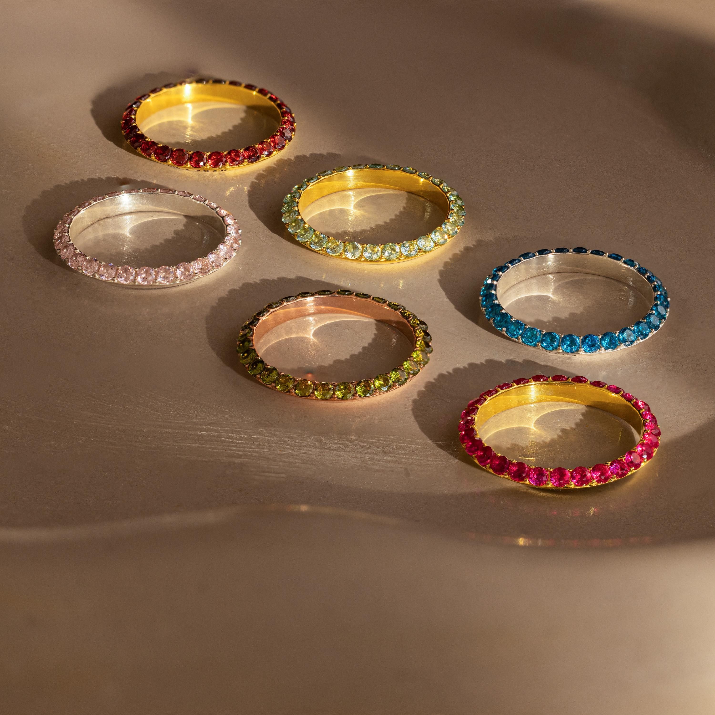 Six colorful gemstone rings—including the Engraved Birthstone Eternity Ring—rest on a smooth, beige surface in soft, warm light.