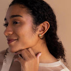 Smiling woman with curly hair rests her chin on her hand, wearing Pave Checkered Huggies against a beige background.