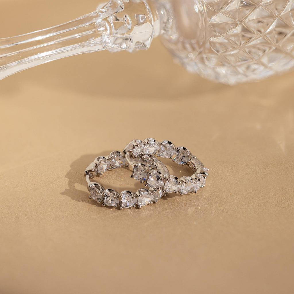 Two silver rings with oval-cut clear gemstones rest on a beige surface beside a crystal glass, paired elegantly with Inside Out Heart Diamond Hoops.