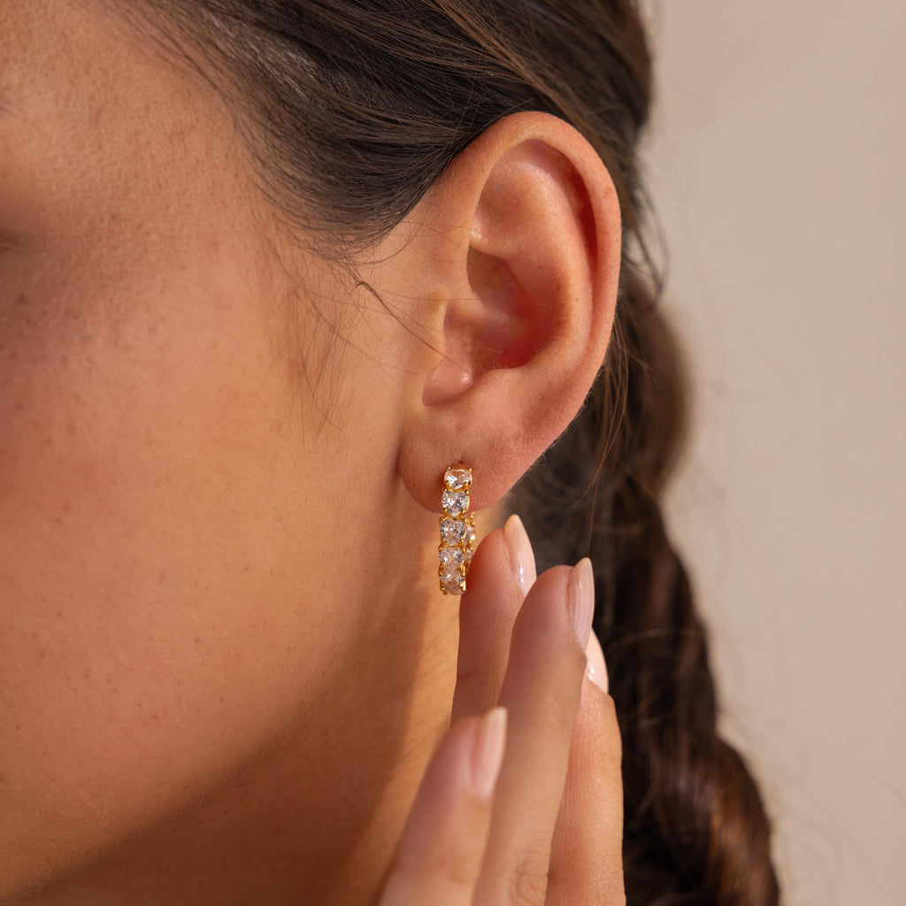 A woman's ear is shown up close wearing the Inside Out Heart Diamond Hoops, featuring small clear gemstones, with her hand near her ear.