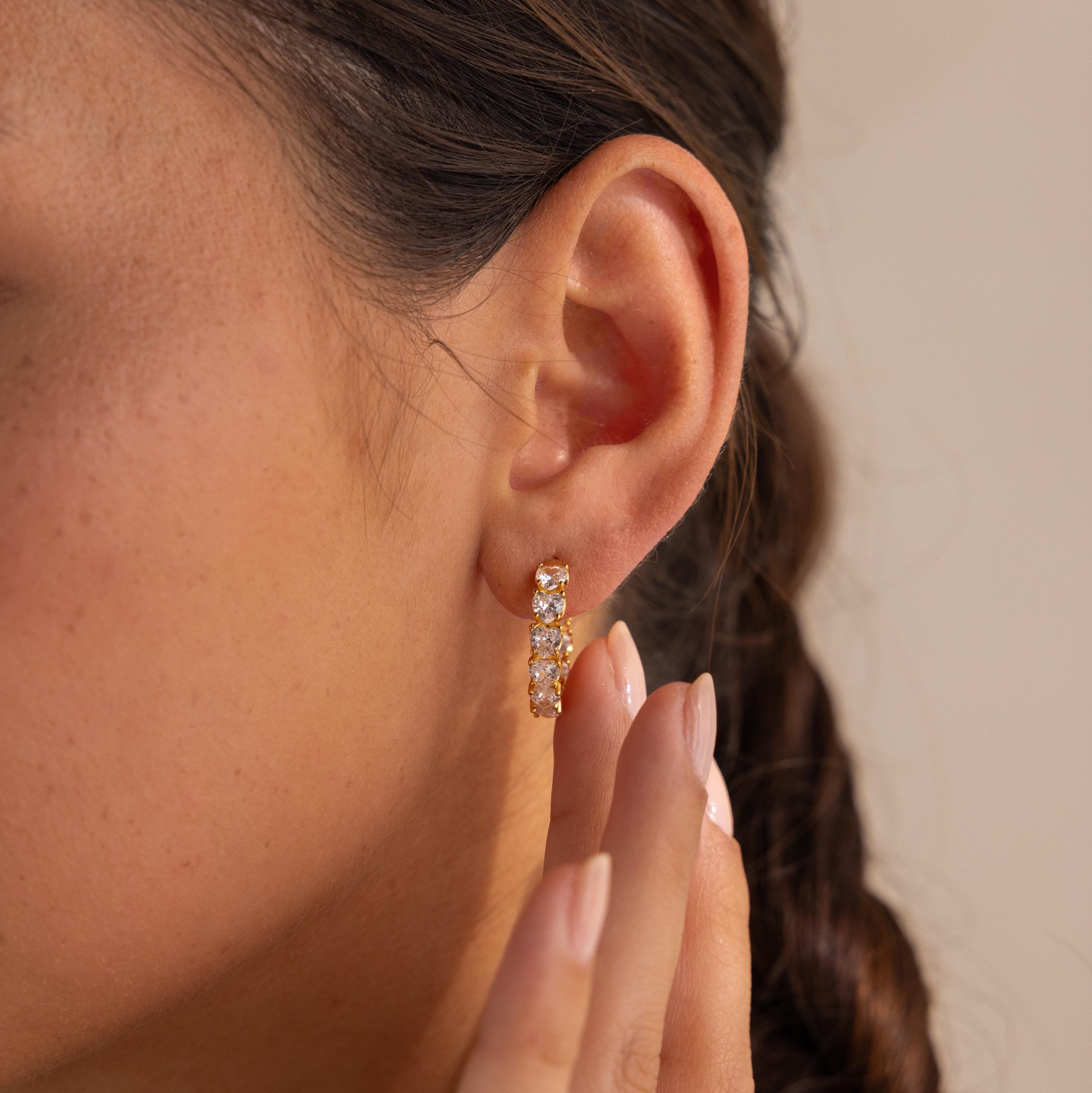 A woman's ear is shown up close wearing the Inside Out Heart Diamond Hoops, featuring small clear gemstones, with her hand near her ear.