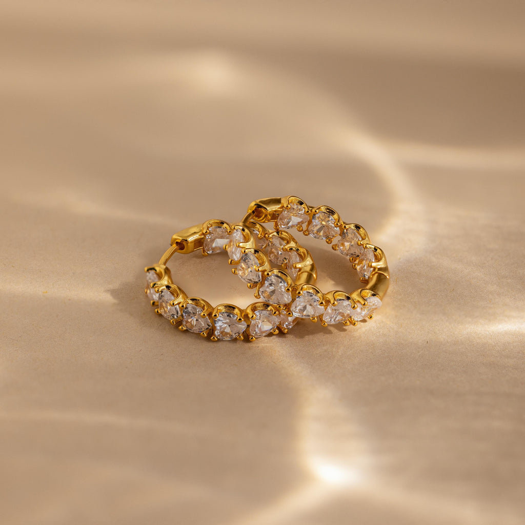 A pair of Inside Out Heart Diamond Hoops, featuring clear gemstones set in gold, displayed on a beige surface with soft light reflections.