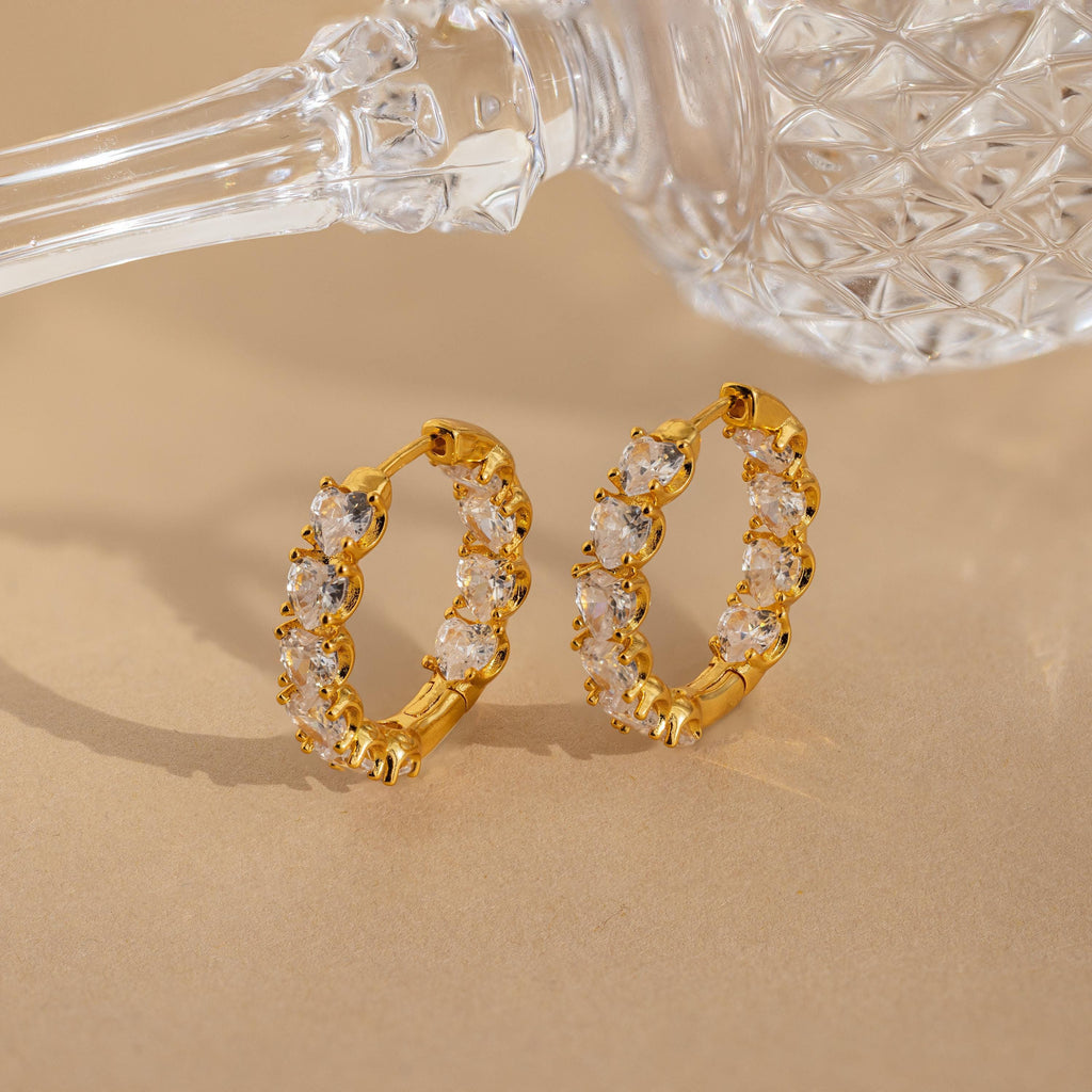 Inside Out Heart Diamond Hoops, gold earrings featuring clear gemstones, displayed next to a faceted glass object on a beige surface.