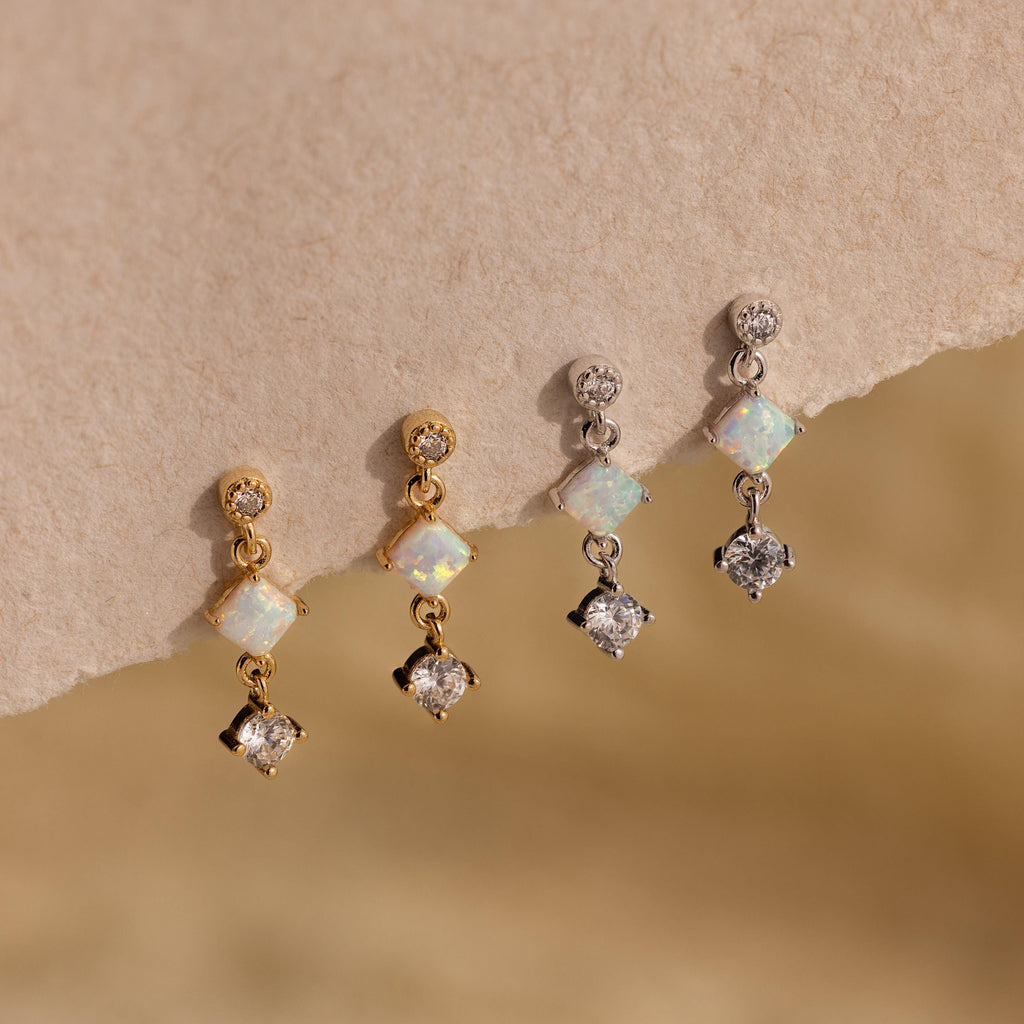 Two pairs of dainty earrings—Opal & Diamond Drop Studs in gold and a silver pair with clear stones—are displayed on a textured beige surface.