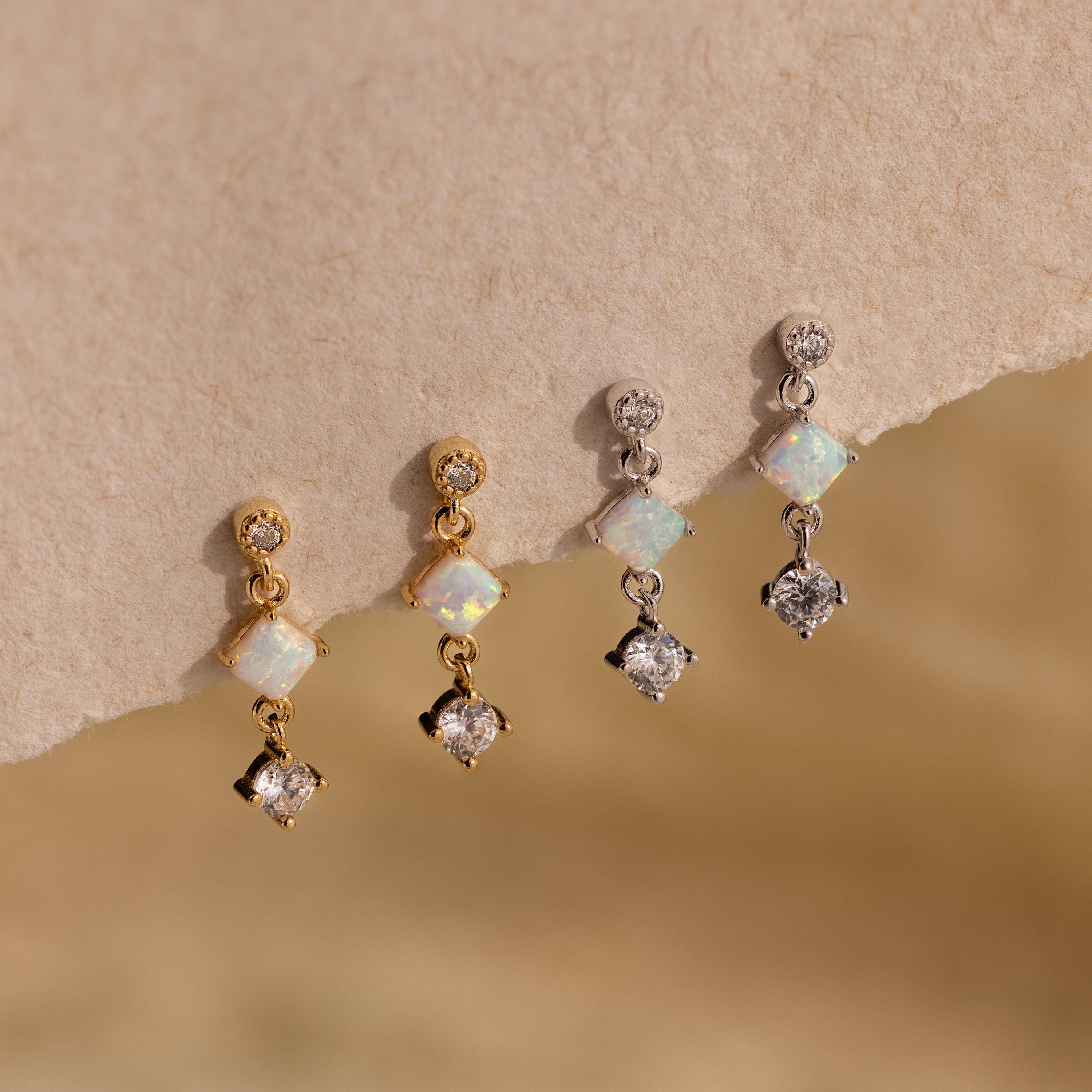 Two pairs of dainty earrings—Opal & Diamond Drop Studs in gold and a silver pair with clear stones—are displayed on a textured beige surface.