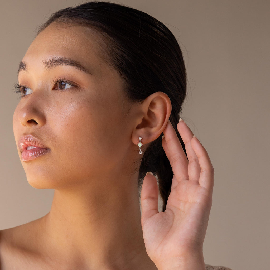 A woman with slicked-back hair touches her ear, highlighting the Opal & Diamond Drop Studs against a neutral background.