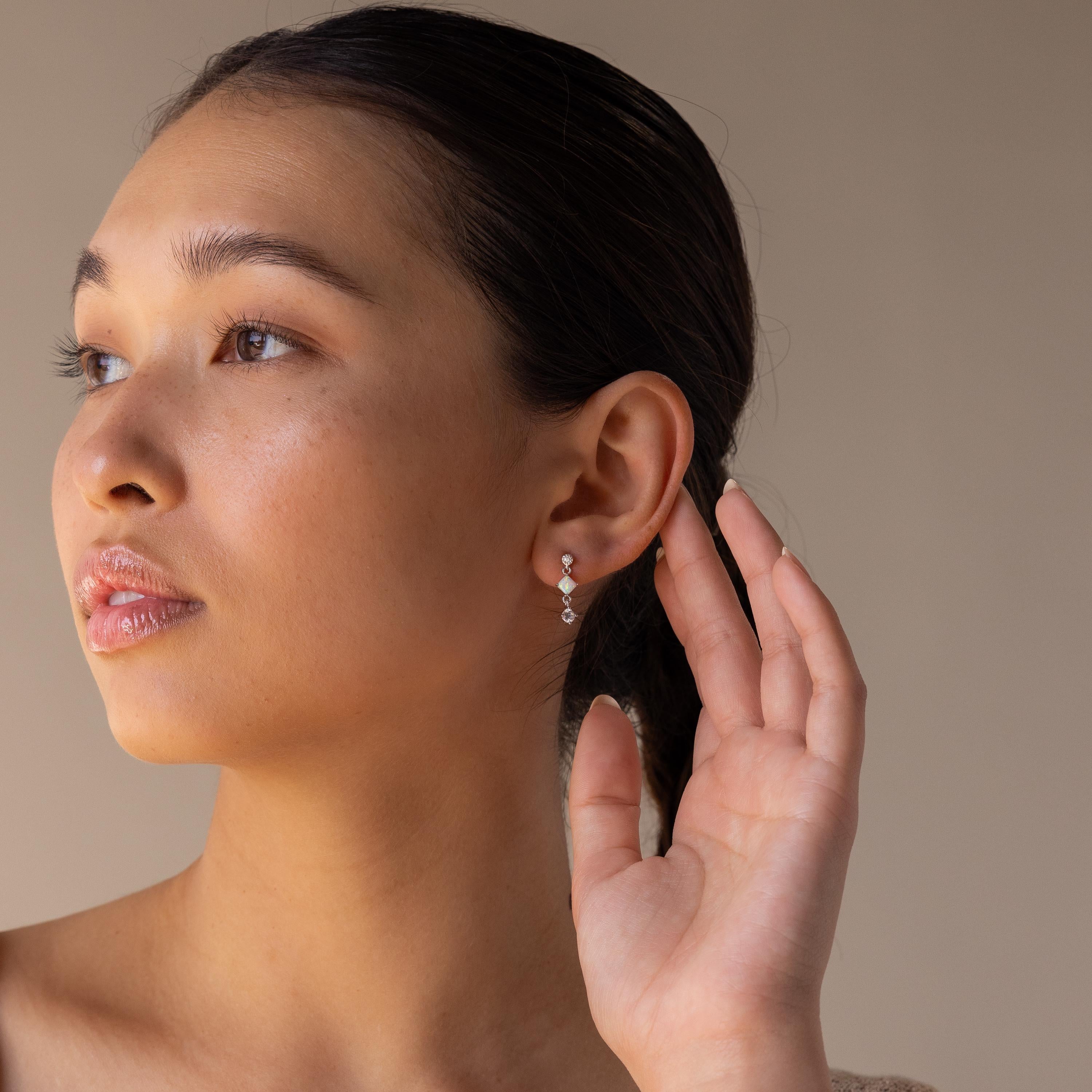 A woman with slicked-back hair touches her ear, highlighting the Opal & Diamond Drop Studs against a neutral background.