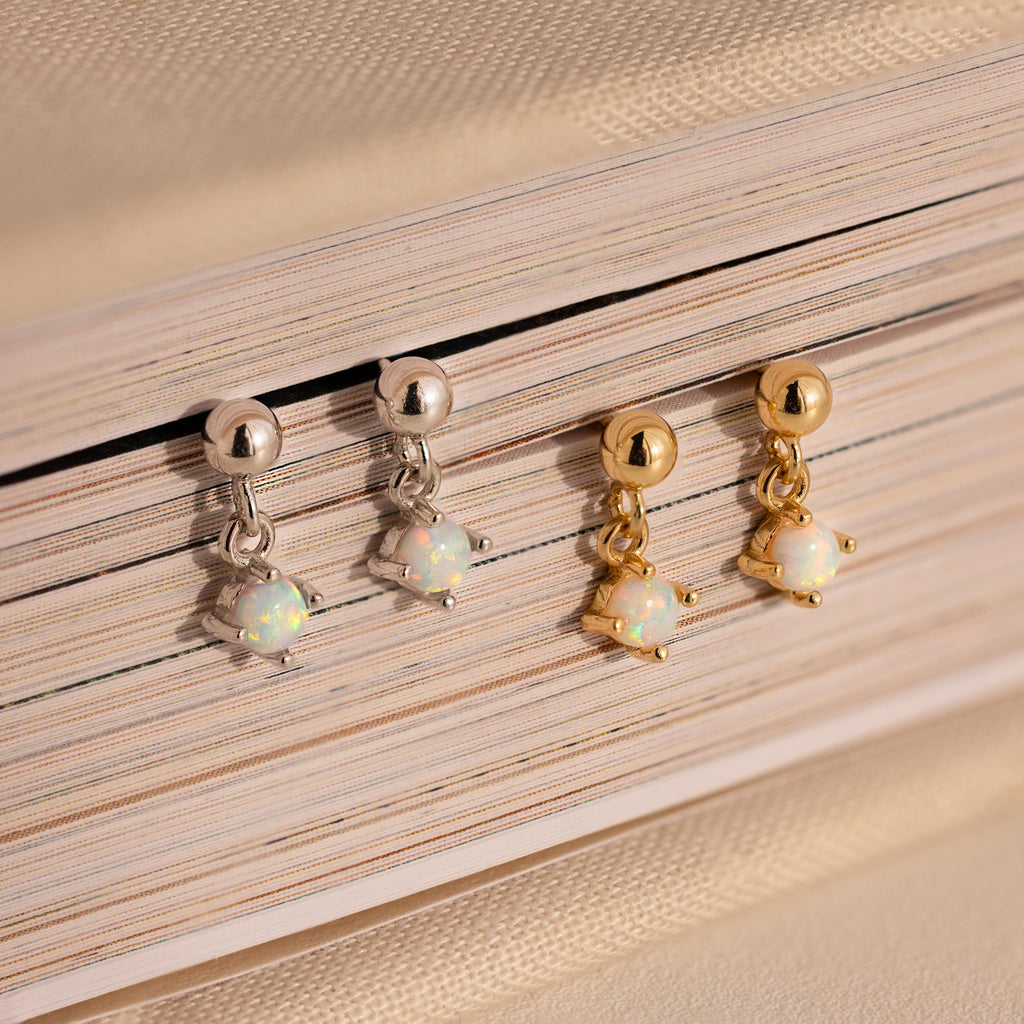 Two pairs of Dainty Opal Drop Studs—one silver and one gold—are elegantly displayed on a closed book, showcasing their lightweight design and adding a touch of sophistication.