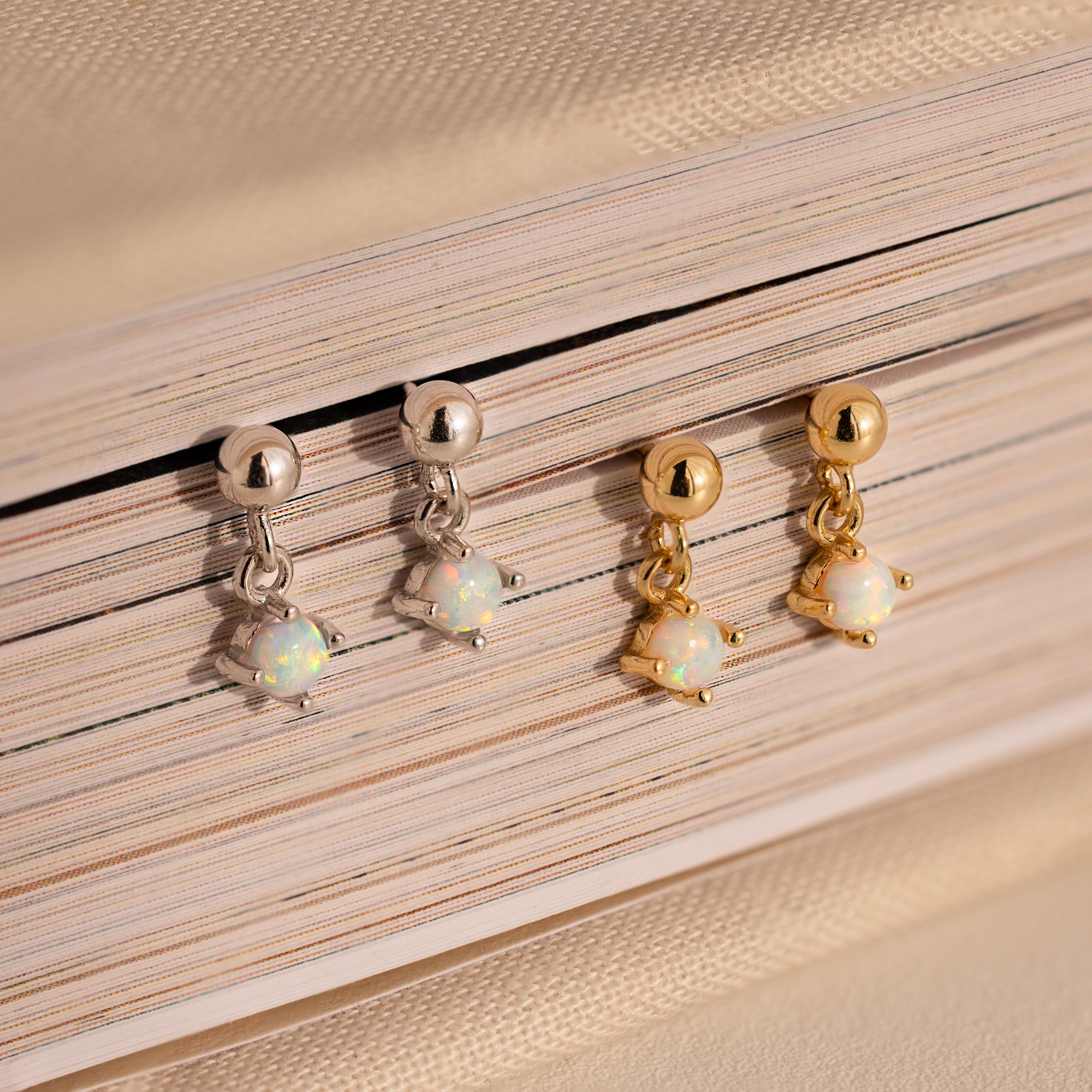 Two pairs of Dainty Opal Drop Studs—one silver and one gold—are elegantly displayed on a closed book, showcasing their lightweight design and adding a touch of sophistication.