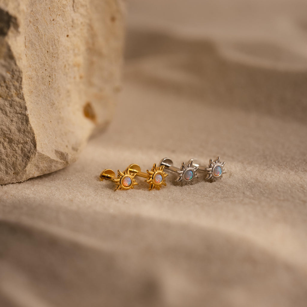 The Opal Sun Flat Back Studs—two silver and one gold—are displayed on sandy ground by a rock, making for the perfect celestial jewelry to elevate your everyday look.