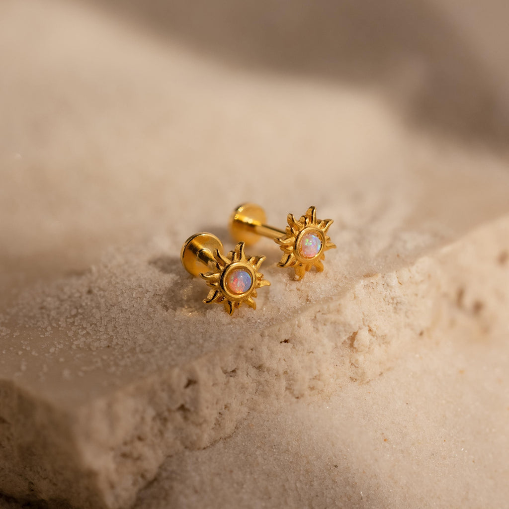 Opal Sun Flat Back Studs feature iridescent centers and a gold finish, bringing celestial style to your look. Shown on a sandy, textured stone surface.