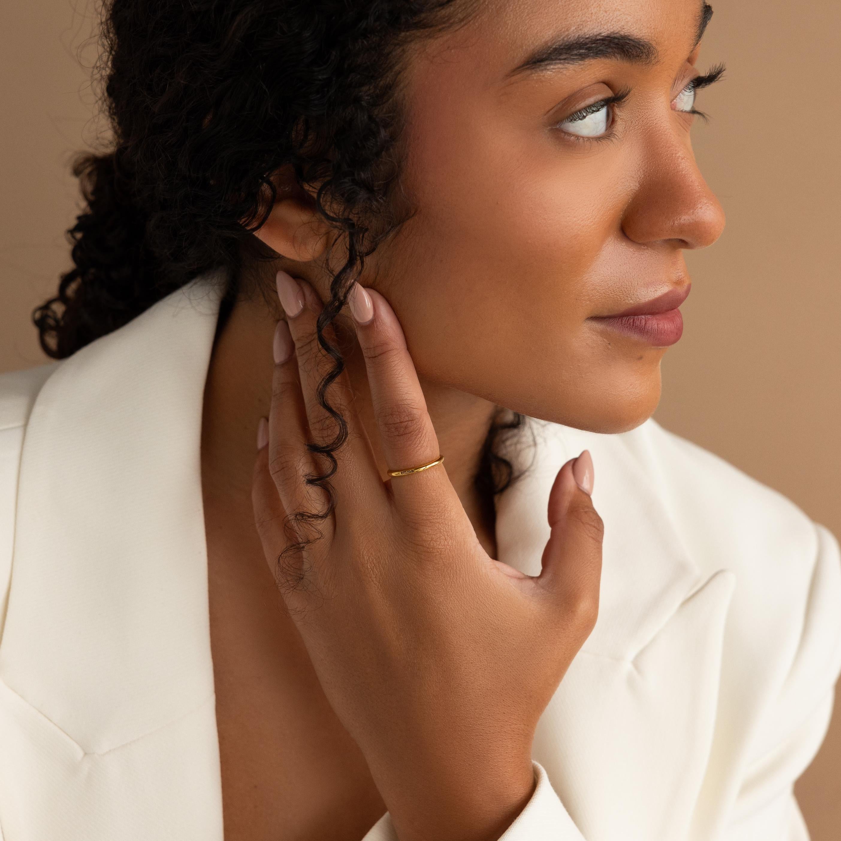 A woman in a white blazer touches her face, highlighting a minimalistic jewelry style with the Engraved Tiny Name Ring, and gazes to the side against a beige background.