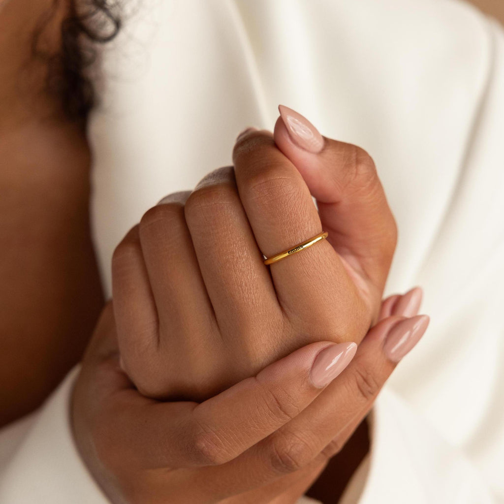 Close-up of nude manicured hands wearing the Engraved Tiny Name Ring, a simple gold piece, against a white fabric background.