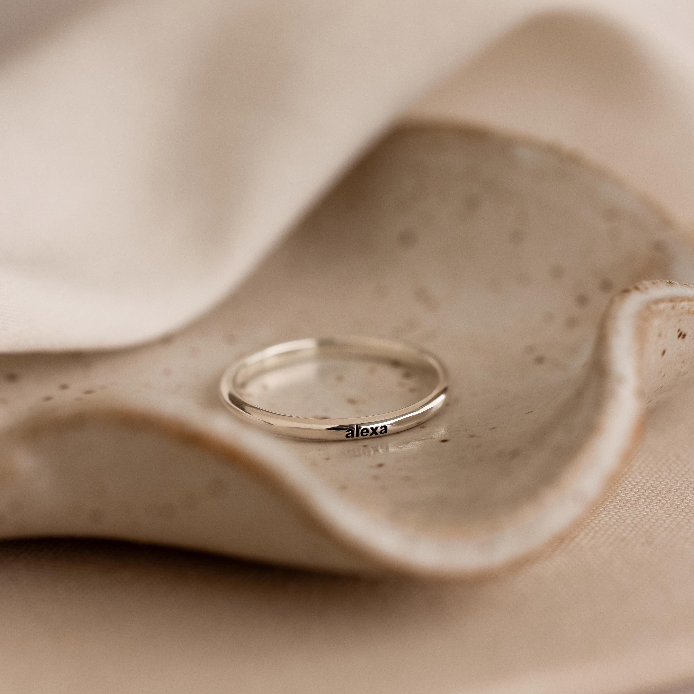 The Engraved Tiny Name Ring is a minimalistic silver band personalized with "alexa," displayed on a textured ceramic dish with soft fabric in the background.