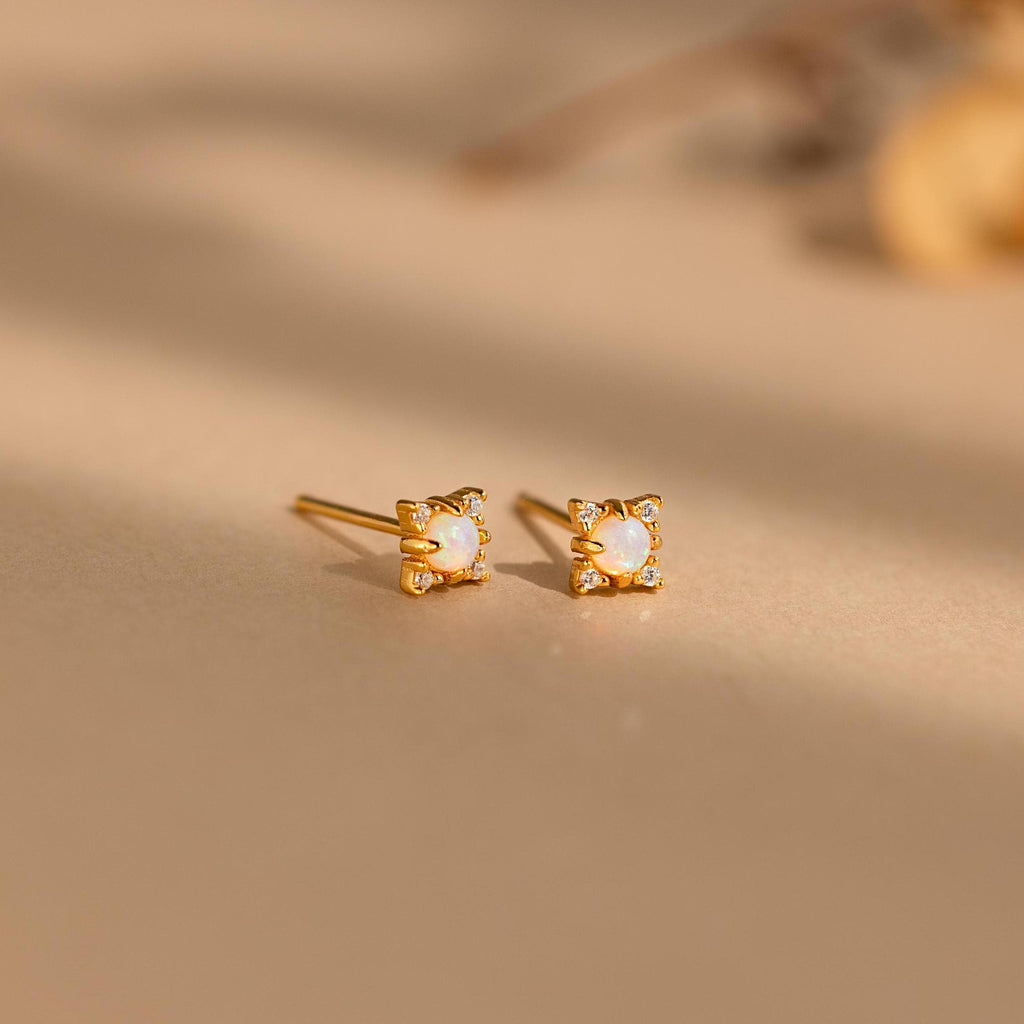 The Opal Diamond Star Studs are small gold earrings with square white stones, inspired by celestial designs and displayed on a beige surface.