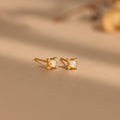The Opal Diamond Star Studs are small gold earrings with square white stones, inspired by celestial designs and displayed on a beige surface.