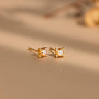The Opal Diamond Star Studs are small gold earrings with square white stones, inspired by celestial designs and displayed on a beige surface.