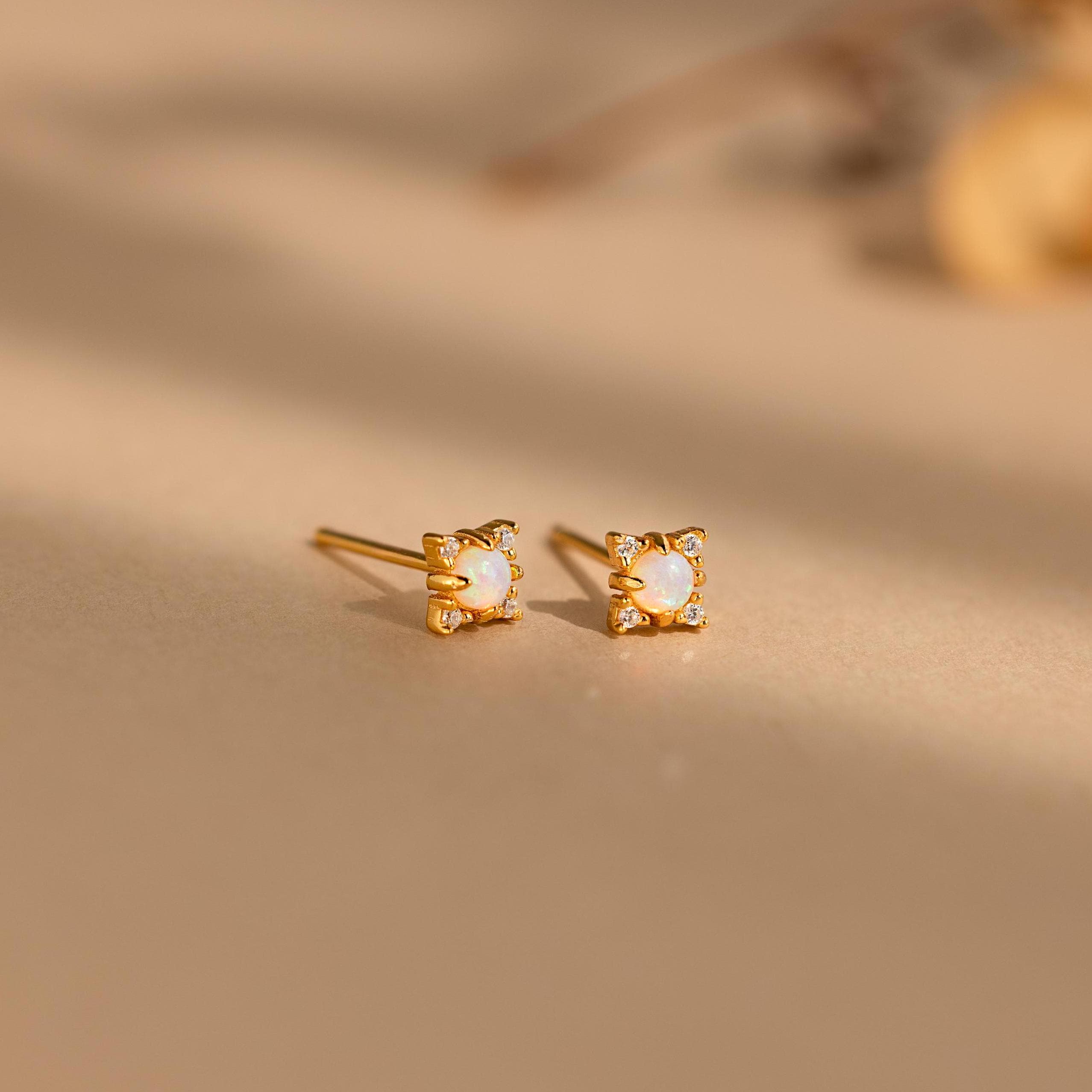 The Opal Diamond Star Studs are small gold earrings with square white stones, inspired by celestial designs and displayed on a beige surface.