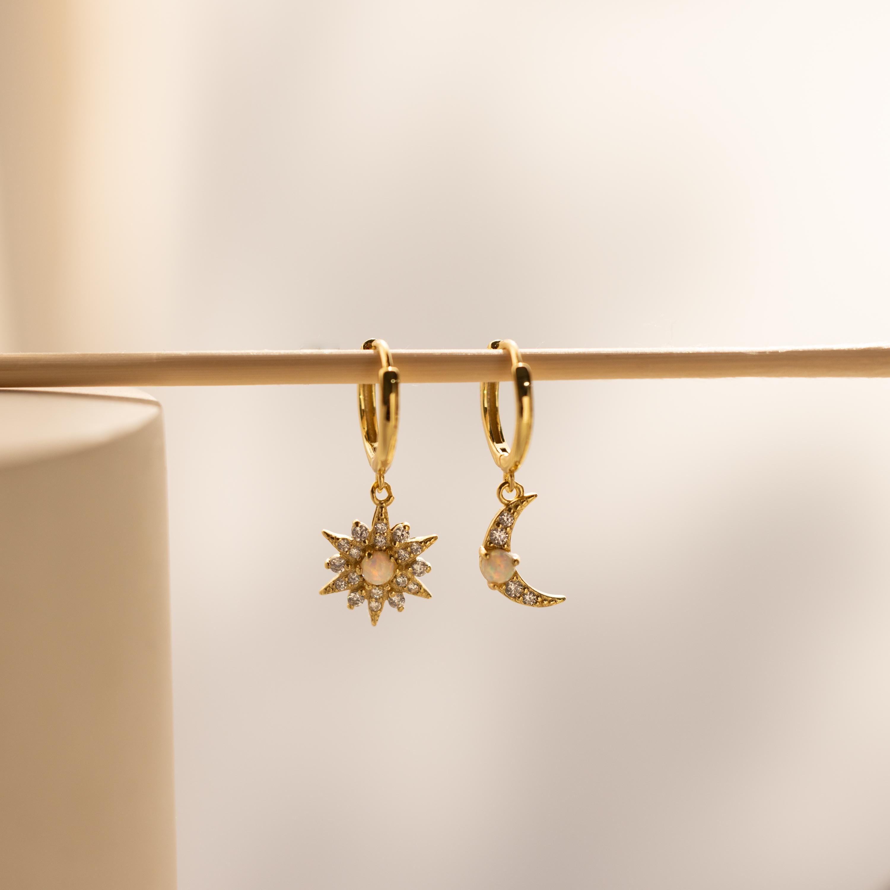 Opal Sun & Moon Huggies are gold hoop earrings with sun and moon charms, each set with small gemstones. Perfect as mismatched cosmic jewelry, they're beautifully displayed hanging on a rod.