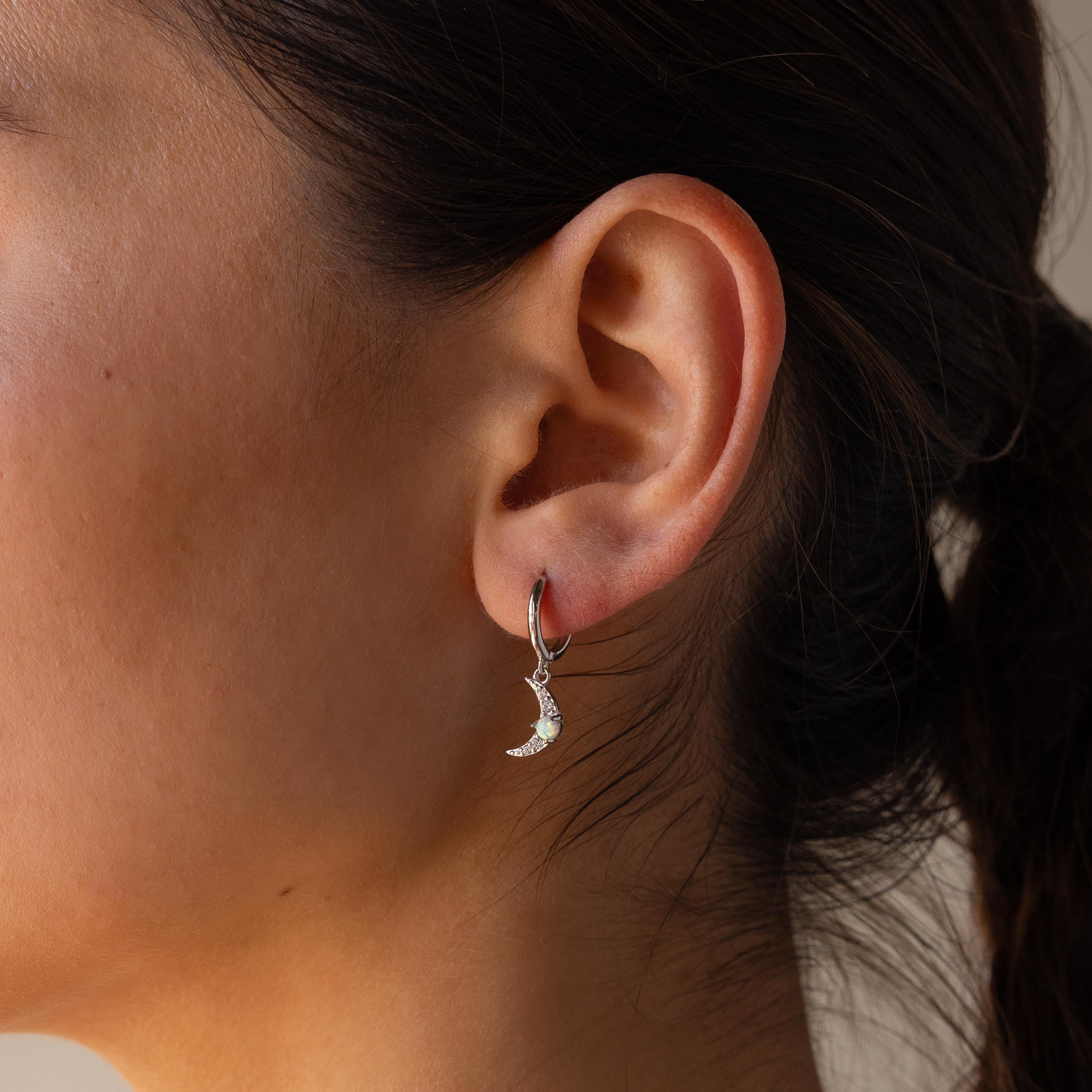 A close-up of a woman’s ear wearing a crescent moon-shaped earring from the Opal Sun & Moon Huggies collection—a delicate silver accessory perfect for fans of celestial jewelry.