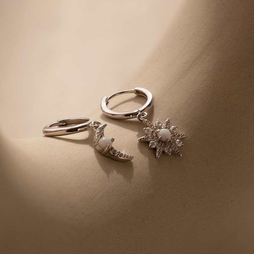 The Opal Sun & Moon Huggies feature one silver earring with a crescent moon charm and another with a starburst, offering cosmic style to any outfit. Displayed on a neutral surface.