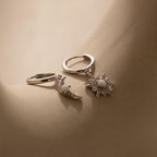 The Opal Sun & Moon Huggies feature one silver earring with a crescent moon charm and another with a starburst, offering cosmic style to any outfit. Displayed on a neutral surface.