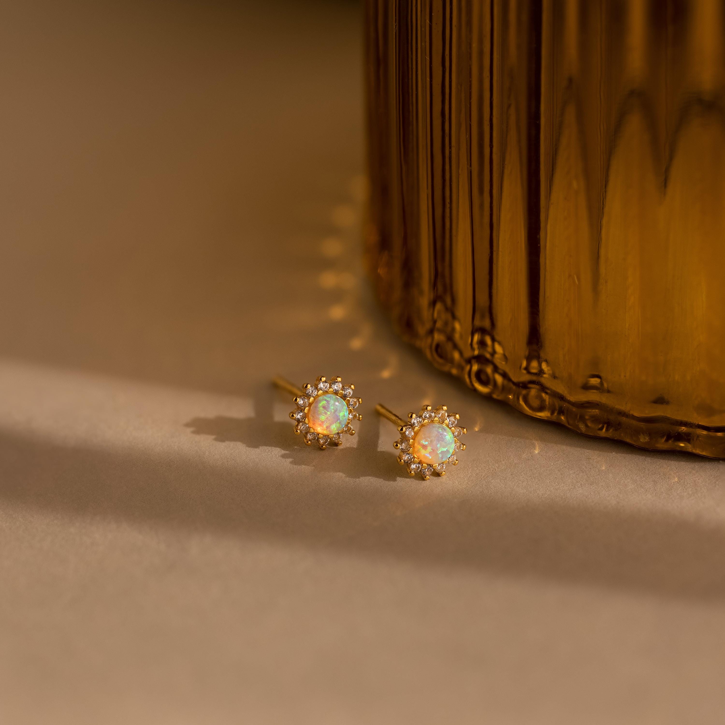 Two Pave Opal Moon Studs and diamond sparkle earrings rest on a beige surface beside a gold, textured glass object, creating an ethereal touch of celestial jewelry.