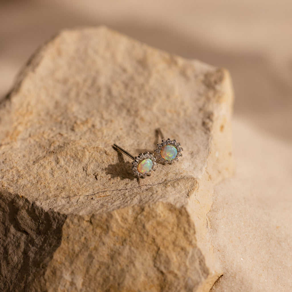 Pave Opal Moon Studs with halo settings rest on a textured beige rock surface in natural light, capturing the essence of celestial jewelry.
