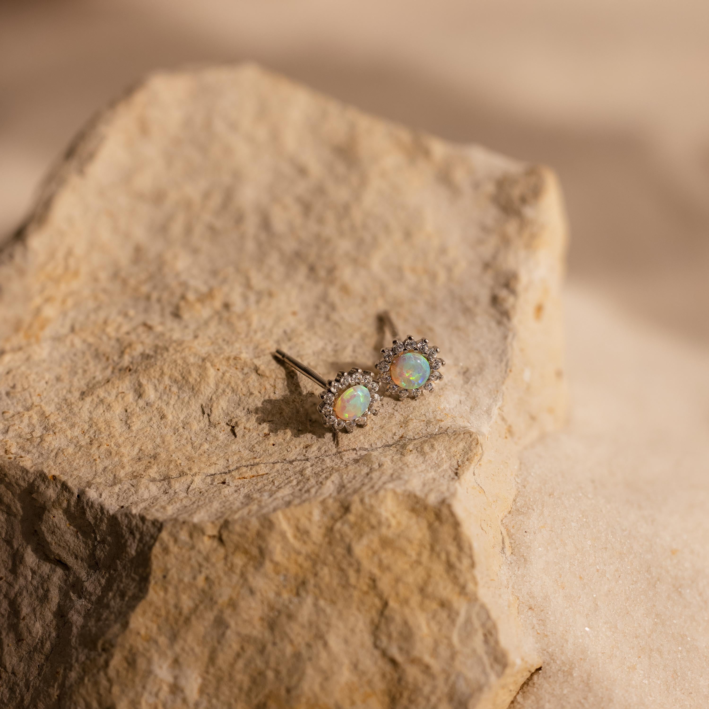 Pave Opal Moon Studs with halo settings rest on a textured beige rock surface in natural light, capturing the essence of celestial jewelry.