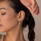 A woman with braided hair wears three gold earrings, including celestial jewelry with sunburst designs and the Pave Opal Moon Studs, adding a diamond-like sparkle to her ear.