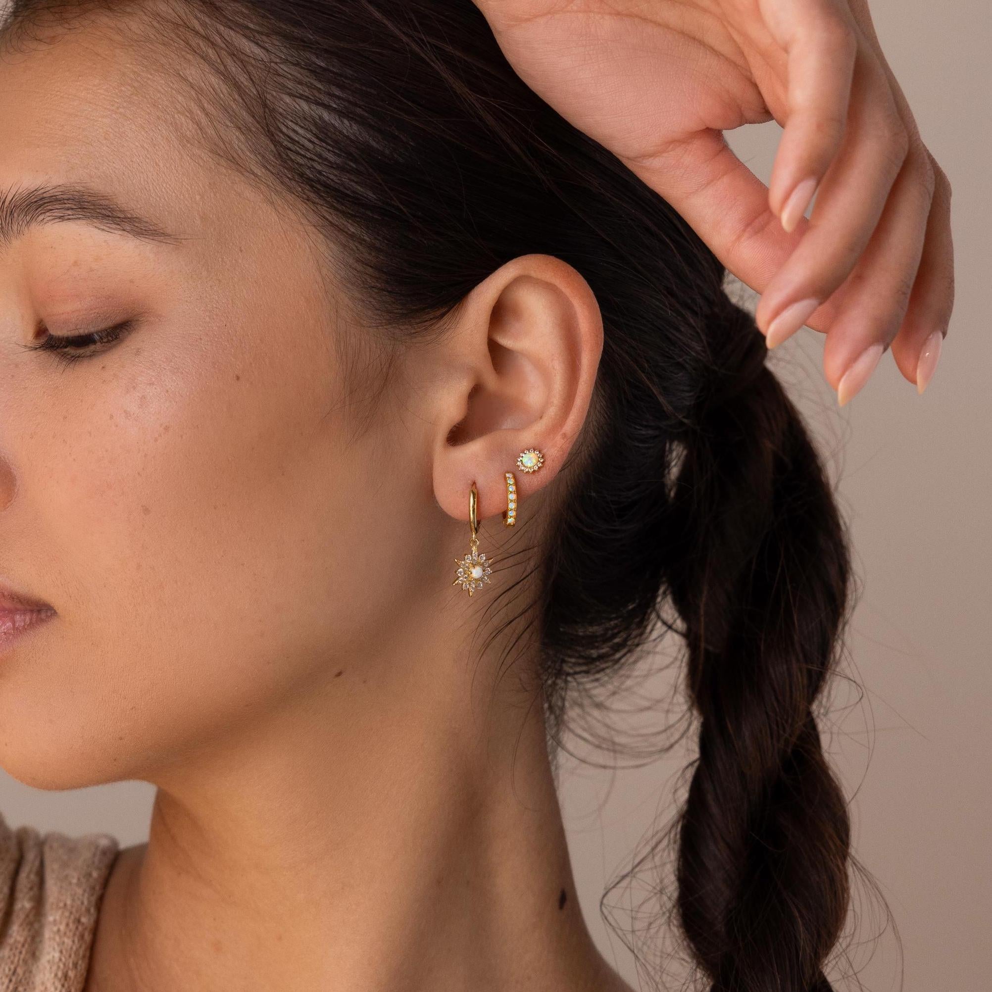 A woman with braided hair wears three gold earrings, including celestial jewelry with sunburst designs and the Pave Opal Moon Studs, adding a diamond-like sparkle to her ear.
