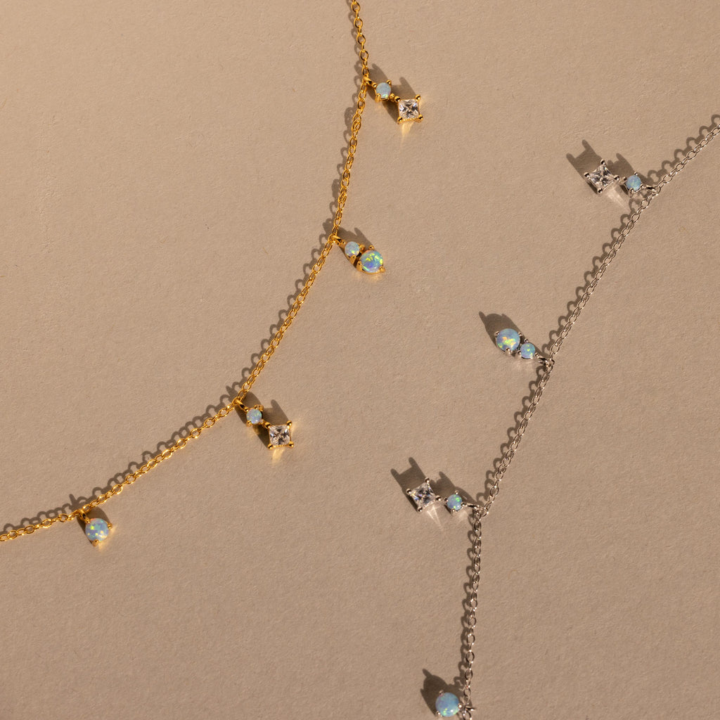 The Opal Diamond Station Necklace set features delicate gold and silver chains with crystal and opal charms, making a perfect bridesmaid keepsake, beautifully displayed on a beige surface.