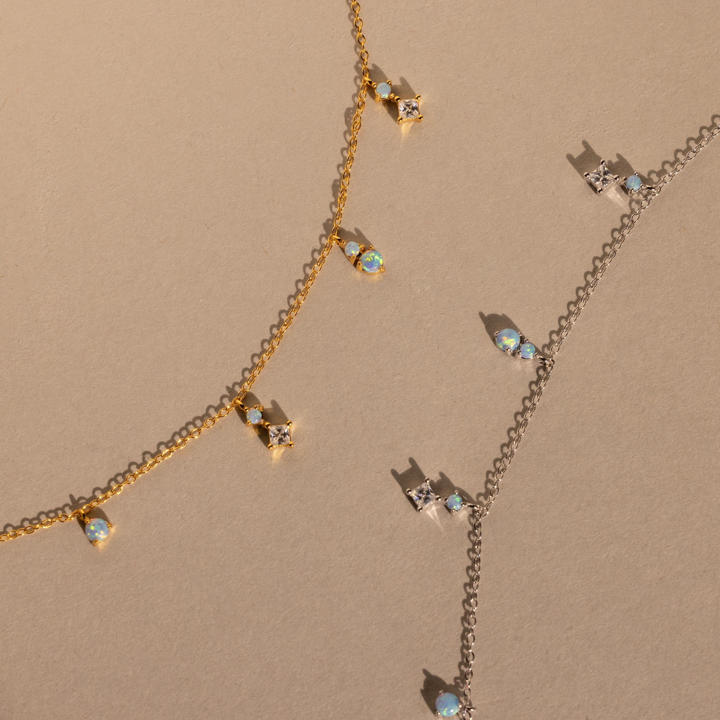 The Opal Diamond Station Necklace set features delicate gold and silver chains with crystal and opal charms, making a perfect bridesmaid keepsake, beautifully displayed on a beige surface.