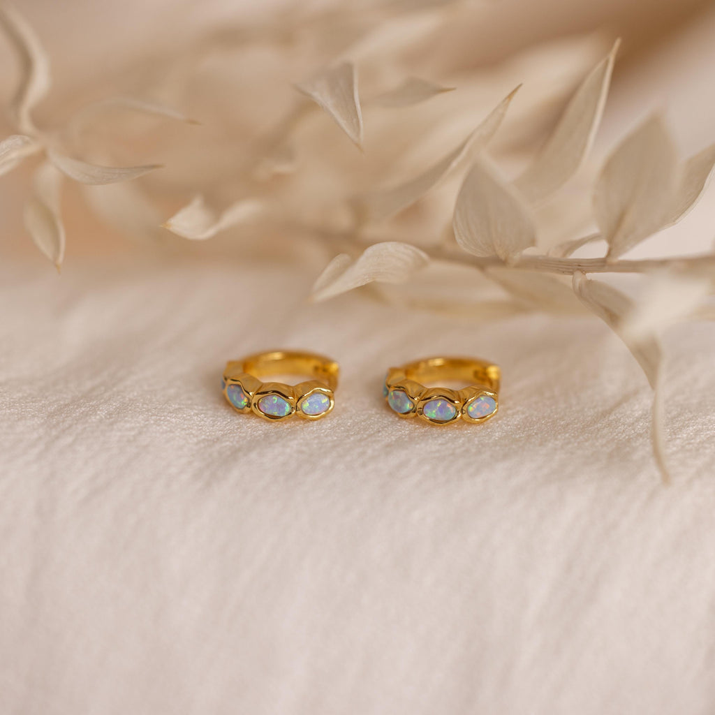 Blue Opal Beaded Huggies rest on soft beige fabric, complemented by dried beige leaves in the background, their gold design and shimmering blue opals recalling the elegance of birthstone jewelry.