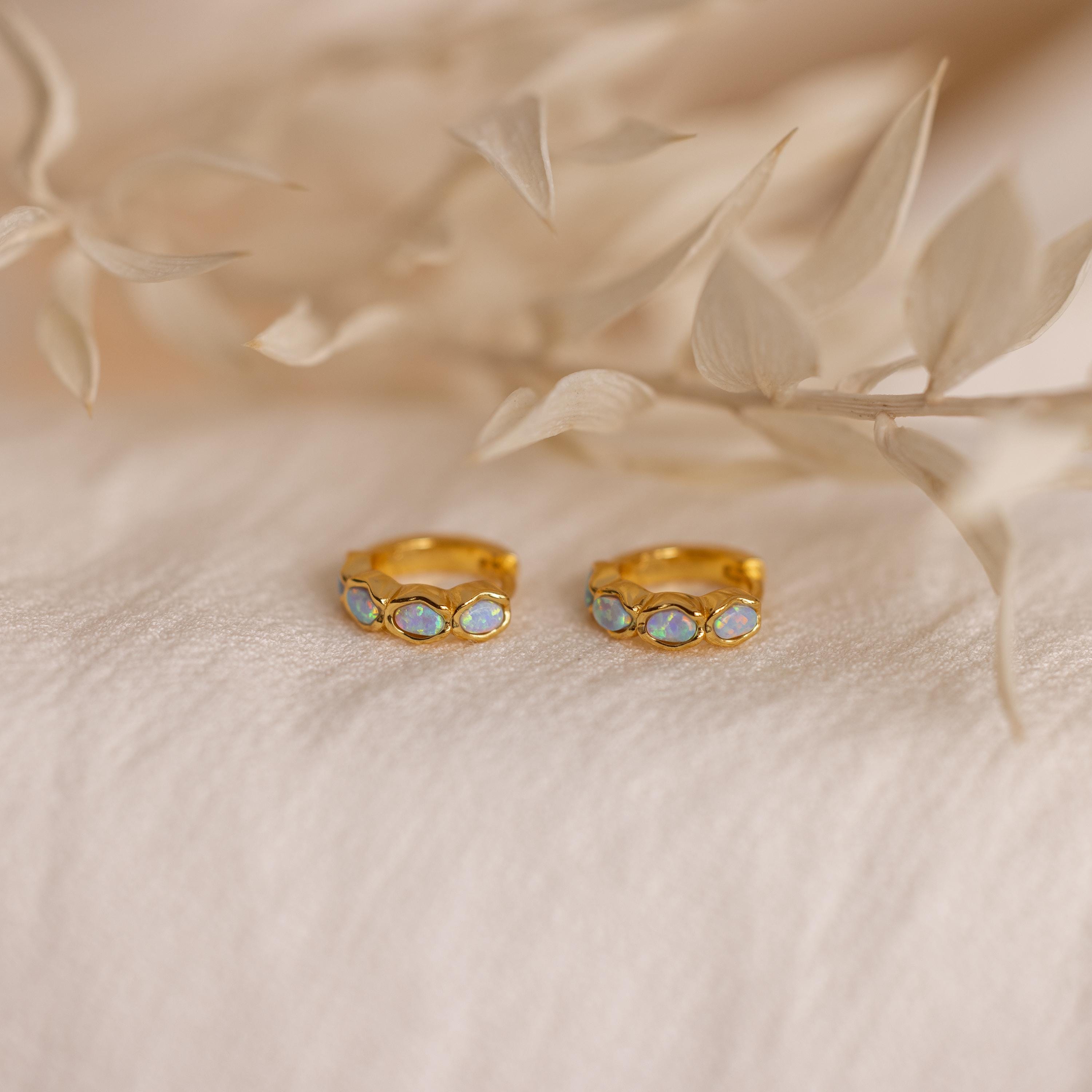 Blue Opal Beaded Huggies rest on soft beige fabric, complemented by dried beige leaves in the background, their gold design and shimmering blue opals recalling the elegance of birthstone jewelry.