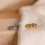 Four hoop earrings—two silver, two gold—feature oval blue opal stones and are displayed on fabric and stone. These elegant Blue Opal Beaded Huggies make stunning birthstone earrings for any jewelry collection.