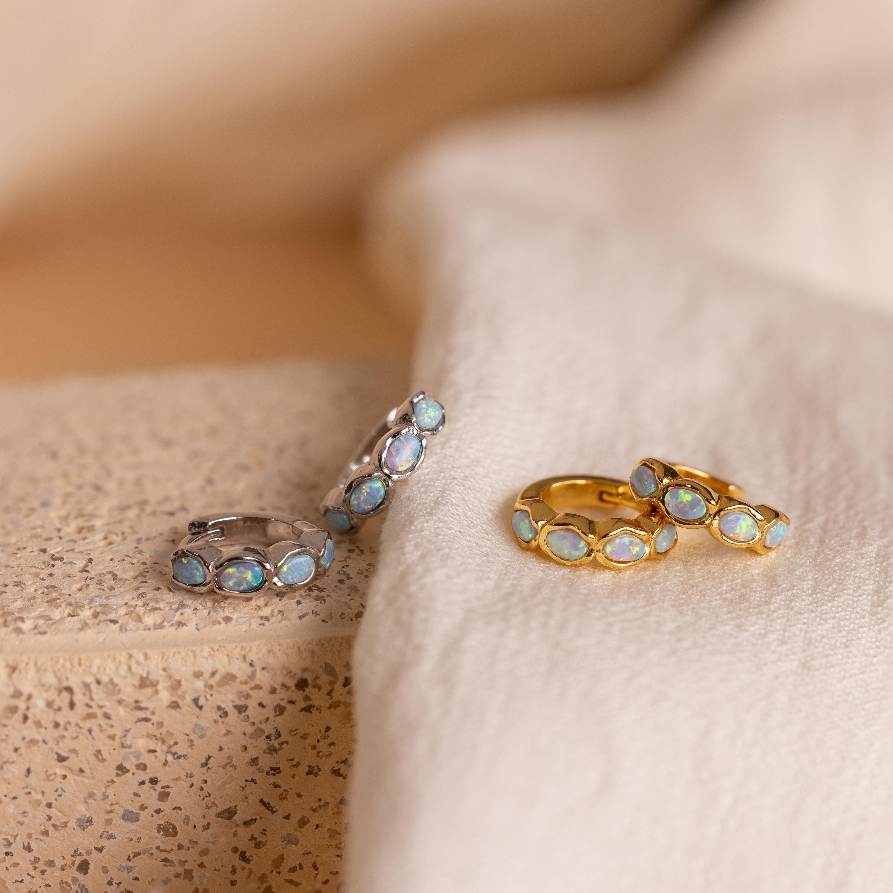 Four hoop earrings—two silver, two gold—feature oval blue opal stones and are displayed on fabric and stone. These elegant Blue Opal Beaded Huggies make stunning birthstone earrings for any jewelry collection.