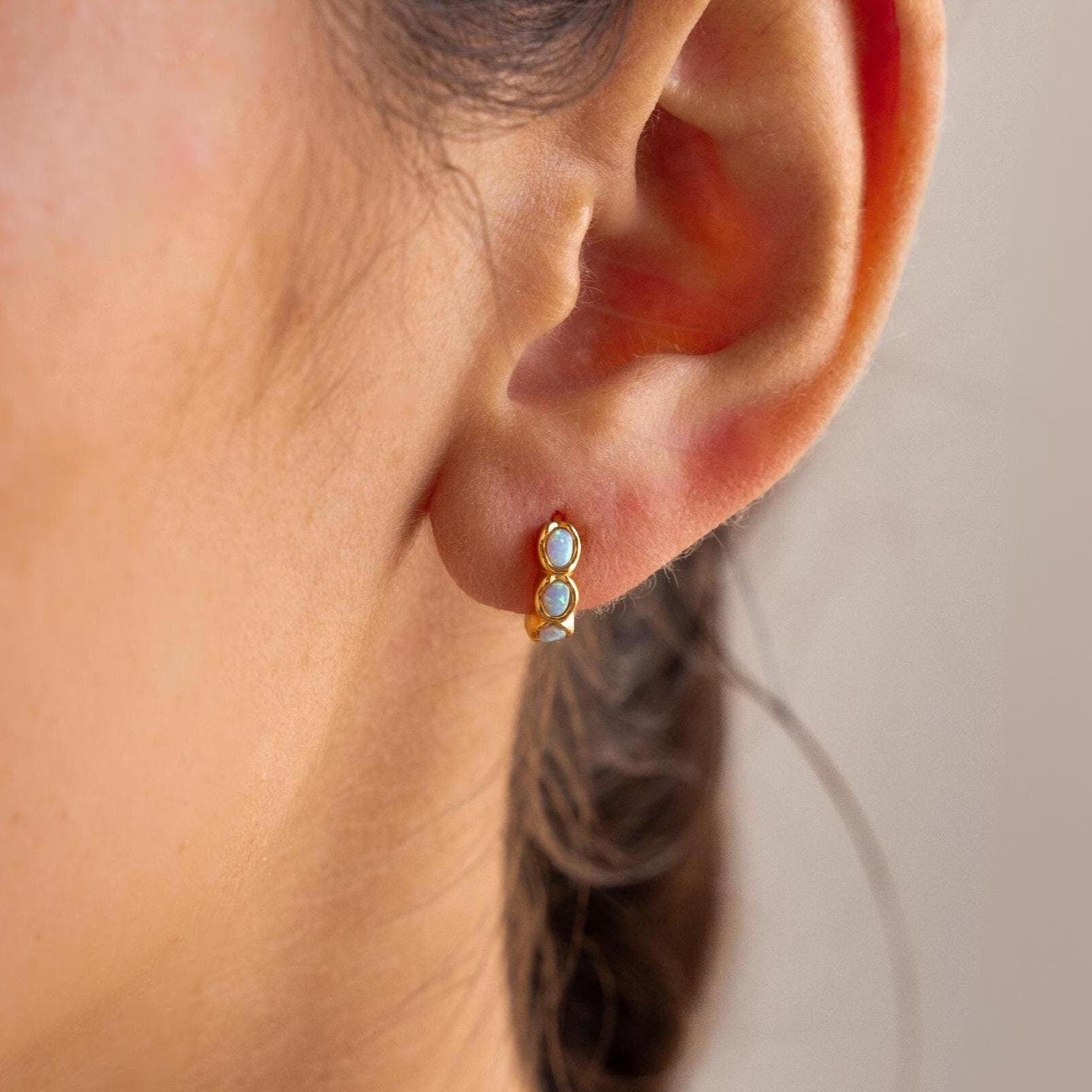 Close-up of an ear wearing the Blue Opal Beaded Huggies, a small gold hoop earring adorned with two light blue opal beads.