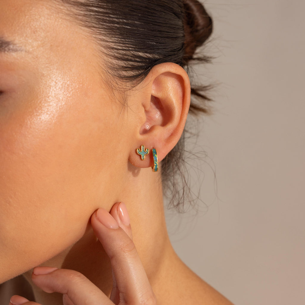 Close-up of a woman’s ear wearing Opal Cactus Studs and a rainbow-shaped earring, her hand resting on her chin.