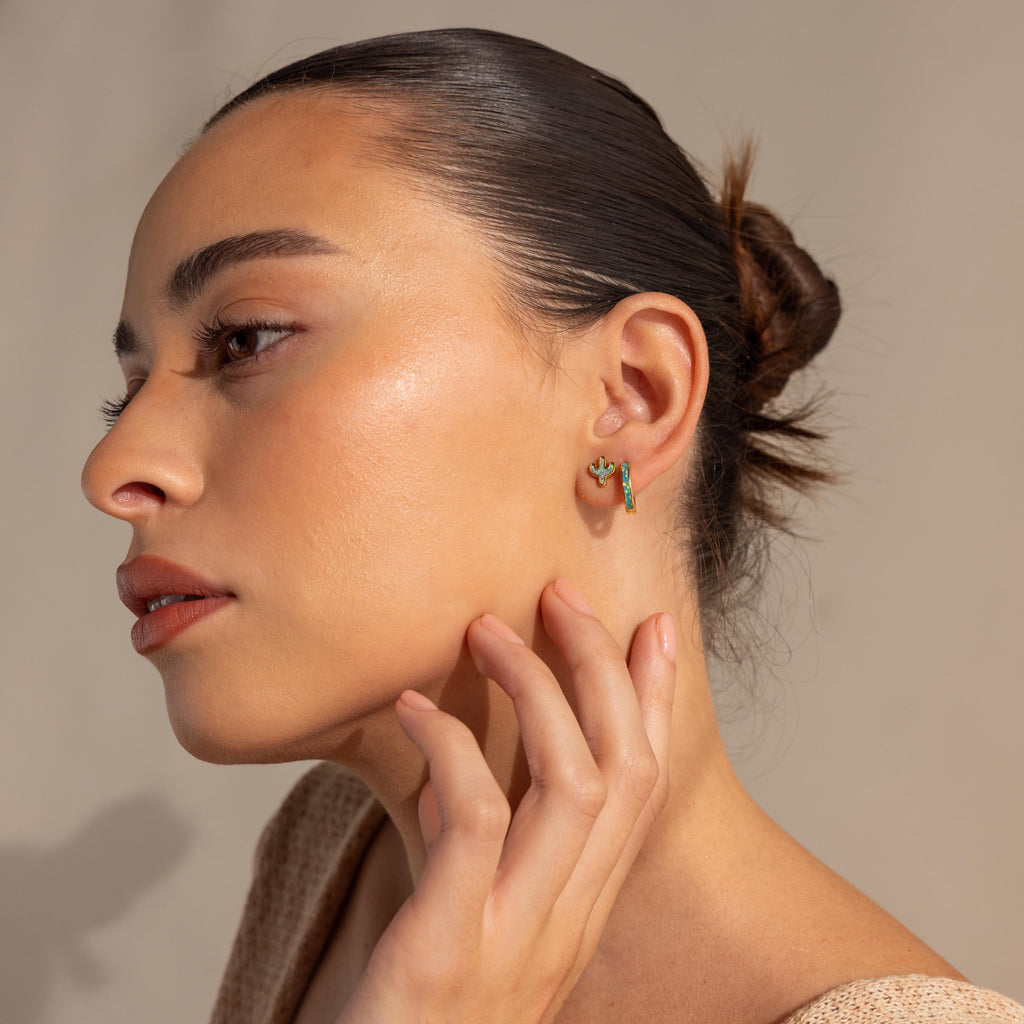 A woman with smooth skin and slicked-back hair touches her ear, highlighting the Opal Cactus Studs—unique earrings that bring a pop of color and charm.