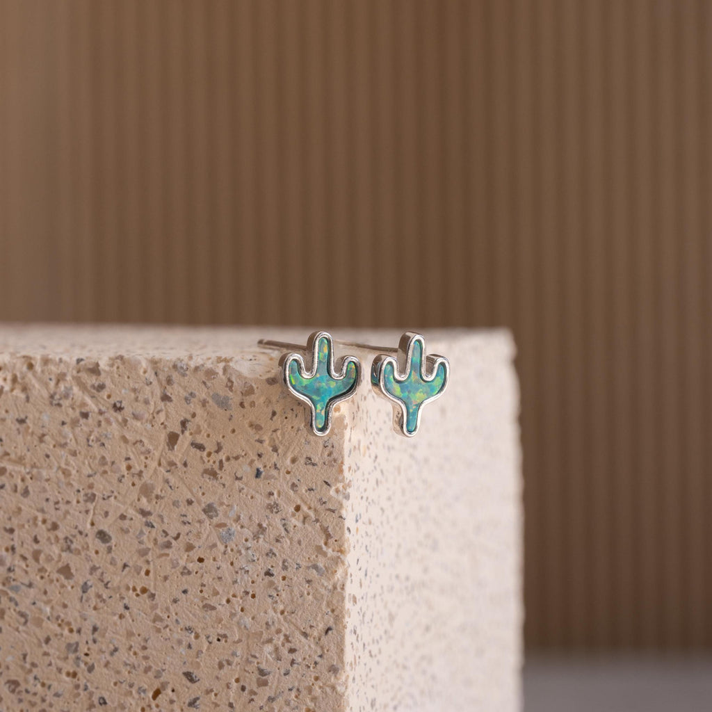 The Opal Cactus Studs are small, green cactus-shaped earrings, shown on a textured stone surface.