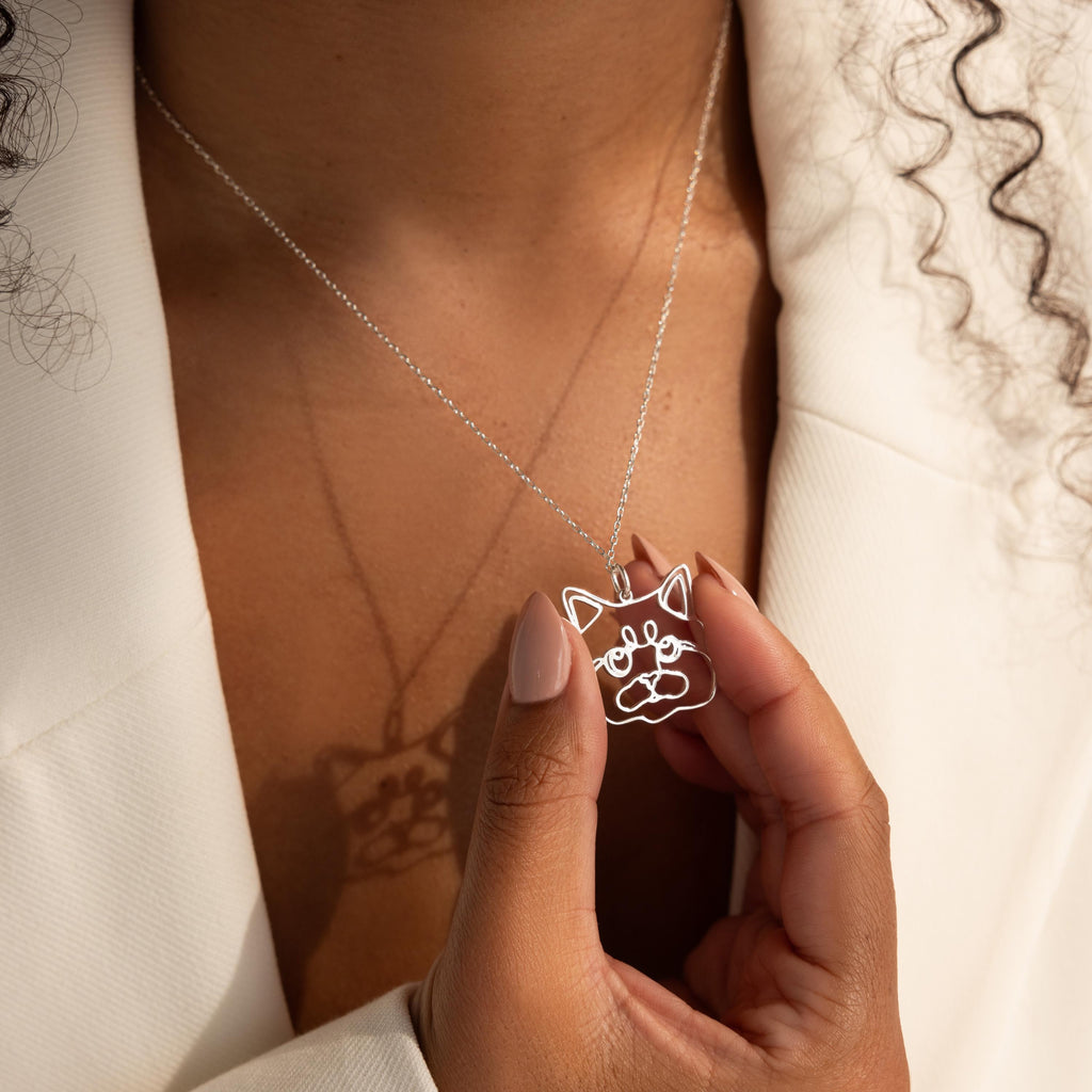 A person wearing a white blazer holds a Custom Pet Line Art Necklace featuring a pendant shaped like a cat’s face.
