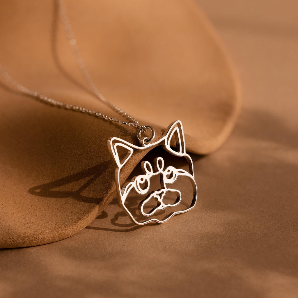 A Custom Pet Line Art Necklace in silver, featuring a cat face pendant, displayed on a brown surface with gentle shadows.