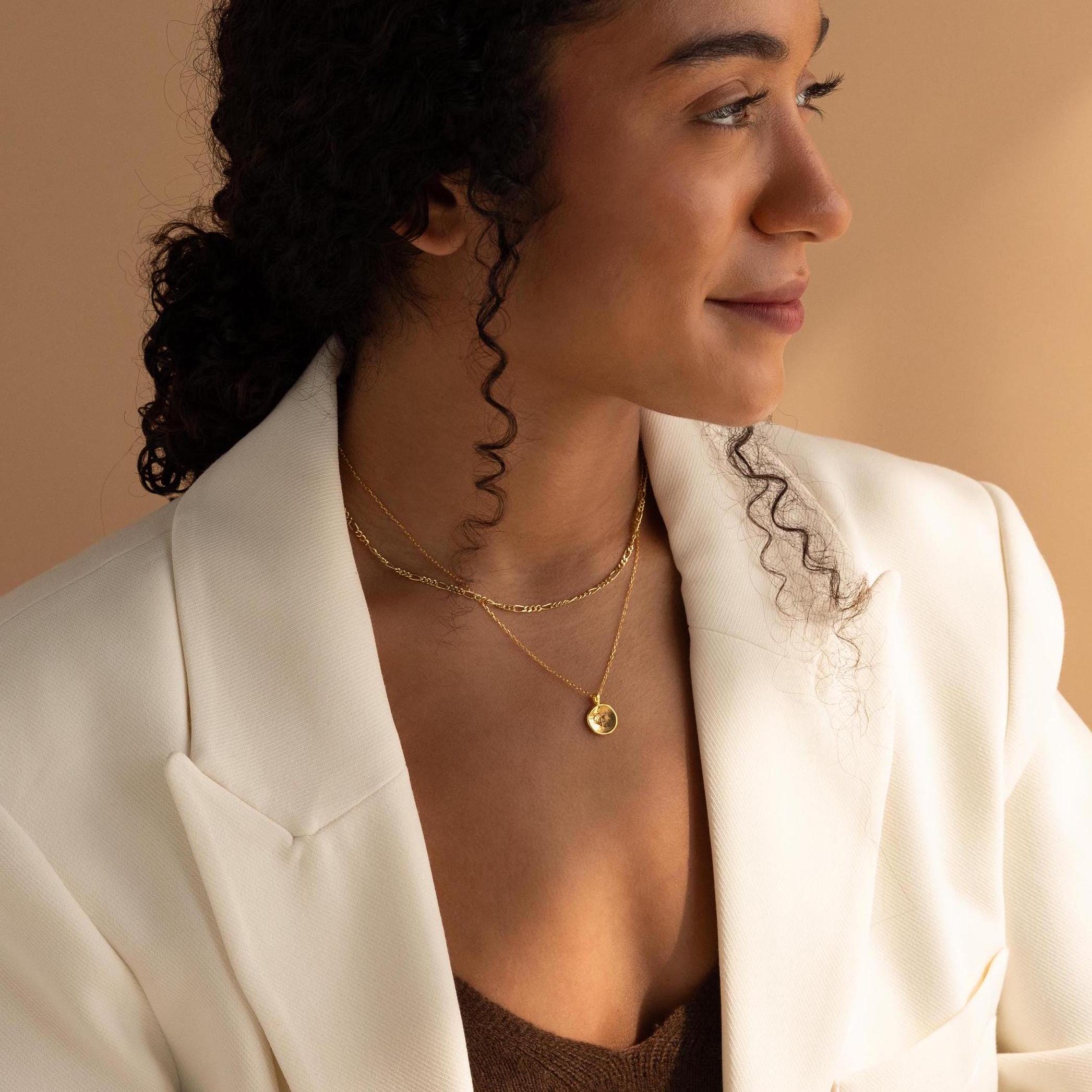 A woman in a white blazer wears layered gold necklaces, one being the Paw Print Coin Necklace, as she looks to the side against a beige background.