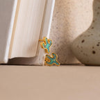 Opal Cactus Studs in gold and green are displayed on the edge of a book, accented by a ceramic vase in the background.