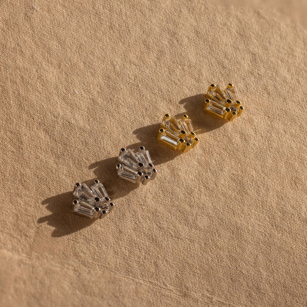 Four pairs of stud earrings, including Art Deco Studs, in silver and gold are displayed on textured beige fabric in natural light.